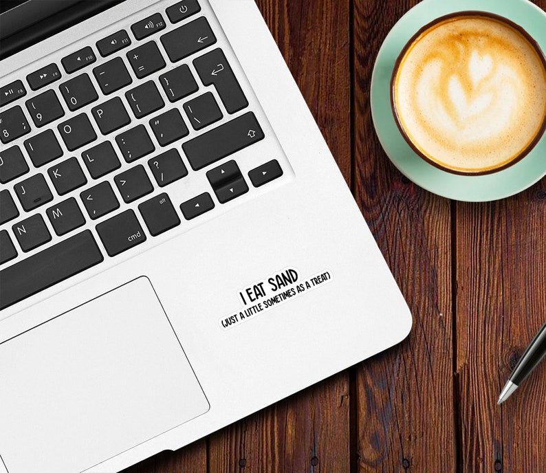 May include: A white laptop computer with a black keyboard and a black trackpad. A sticker on the laptop reads "I eat sand (just a little sometimes as a treat)".