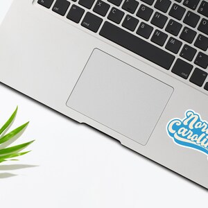 K&ouml;nnte beinhalten: Ein blauer und wei&szlig;er Aufkleber mit dem Text "North Carolina" in einer Retro-Schriftart, platziert auf einem silbernen Laptop.
