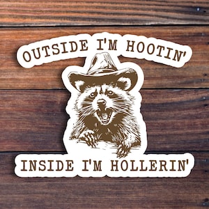 May include: A white sticker with a raccoon wearing a cowboy hat. The sticker says "Outside I'm Hootin' Inside I'm Hollerin'".