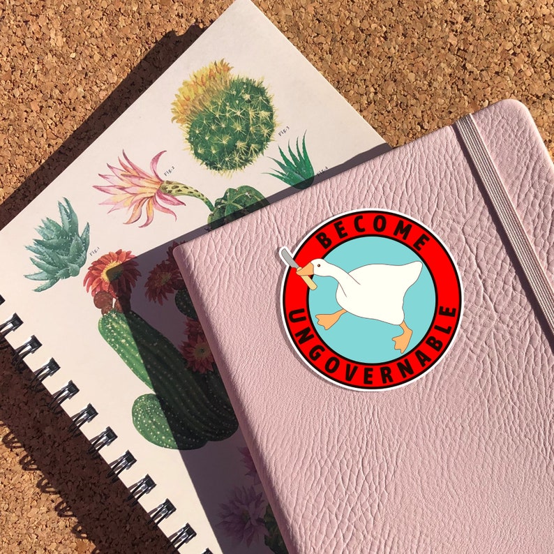 Peut inclure: Un carnet rose avec un autocollant blanc et rouge qui dit "Become Ungovernable" avec une oie de dessin anim&eacute; tenant un couteau.