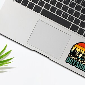 May include: A vintage-style sticker with a sunset background and a silhouette of trees. The sticker says "I love peeing outside". The sticker is on a silver laptop.