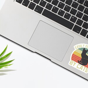 May include: A vintage-style sticker with a black cat silhouette and the text "Easily Distracted By Cats". The sticker has a red, orange, and yellow color scheme.