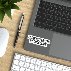 May include: A white laptop computer with a black keyboard and trackpad. A sticker on the trackpad reads "EASILY DISTRACTED BY CHOCOLATE". A white wireless mouse, a black pen, and a green plant are on the wooden desk surface.