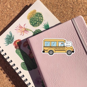 May include: A yellow school bus sticker with a white goose driving. The bus says "STRUGGLE BUS" and the goose has a speech bubble that says "Get in, bestie!"