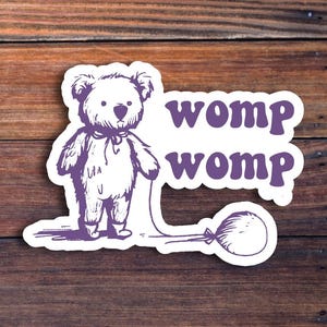 May include: A white sticker with a cartoon teddy bear holding a balloon. The text "womp womp" is printed in purple on the sticker.