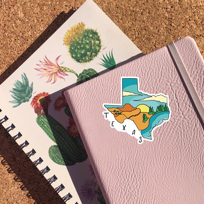 May include: A pink notebook with a colorful sticker of the state of Texas with the word "TEXAS" written on it. The sticker depicts a desert landscape with a sunset and a lake.