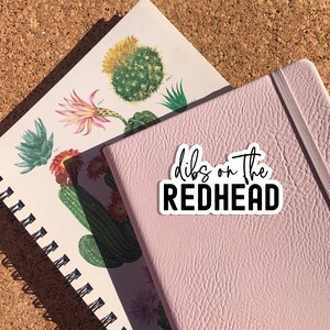 May include: A pink notebook with a white sticker that says "dibs on the redhead" in black cursive lettering.