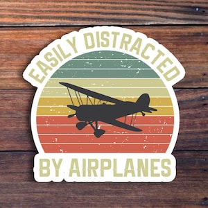May include: A vintage-style sticker with a red, orange, and yellow sunset background. The sticker says "Easily Distracted by Airplanes" with a silhouette of a biplane in the center.