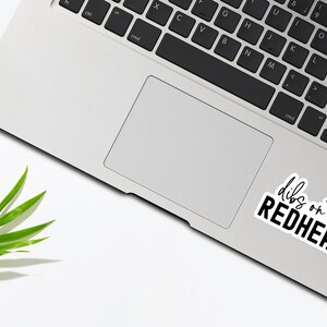 May include: A laptop computer with a sticker that says "dibs on the redhead" on the trackpad. A green leaf is in the lower left corner of the image.
