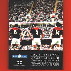 May include: A program cover for the RBS 6 Nations Wales vs. England rugby match. The image features three rugby players in black and red jerseys with numbers 8, 4, and 14, standing on a field in front of a packed stadium. The cover includes text and logos.
