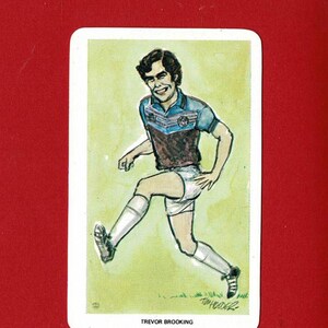 May include: A vintage football trading card showing a cartoon illustration of Trevor Brooking in a maroon and blue kit, white shorts, and white socks. The player is kicking a ball. The card is set against a red background.
