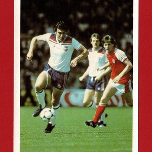 May include: A vintage football trading card showing a player in a white shirt with the England flag on the sleeve, dribbling the ball down the pitch. The player is wearing blue shorts and white socks. Two other players are in the background, one in a white shirt and the other in a red shirt.