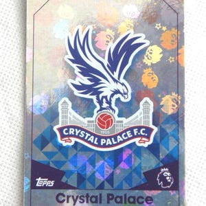 Può includere: Carta collezionabile del Crystal Palace F.C. con il logo della squadra: un'aquila blu e bianca sopra un pallone da calcio rosso e il nome della squadra. La carta ha uno sfondo olografico e il logo Topps.