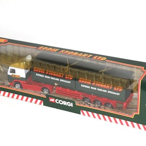 May include: A Corgi model truck in a box. The truck is red, white, and green, with the words "EDDIE STOBART LTD" and "EXPRESS ROAD HAULAGE SPECIALIST" printed on the side. The box is green with red and white stripes.