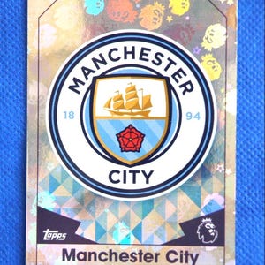 Puede incluir: Tarjeta coleccionable del Manchester City con el escudo del equipo, un escudo con un barco, una rosa roja y las palabras "Manchester City". La tarjeta tiene un fondo holográfico y el logotipo de Topps.