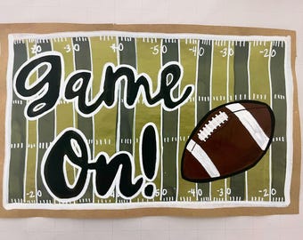 May include: A hand-painted sign with the words "Game On!" in black lettering on a green and brown football field background. A brown football with white laces is in the lower right corner.