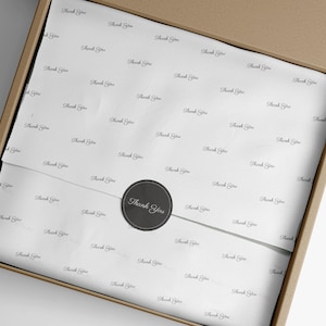 May include: A brown cardboard shipping box containing white tissue paper printed with the repeating black text Thank You. The tissue paper is secured in the center by a circular black sticker featuring the white text Thank You in script.