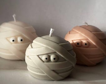 May include: Three mummy-shaped candles with googly eyes. The candles are white, light brown, and gray.