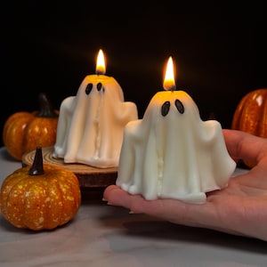Puede incluir: Dos velas encendidas con forma de fantasma, con ojos negros y apariencia de sábana blanca. Pequeñas calabazas naranjas rodean las velas, creando un tema de Halloween. Las llamas son de color amarillo brillante.