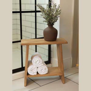 May include: A wooden stool with a shelf, featuring a natural wood finish. The stool has a simple, minimalist design with a rectangular top and angled legs. A vase with dried greenery sits on the top shelf, and white towels are neatly stacked on the lower shelf.