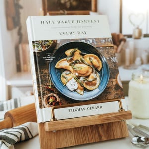 Könnte beinhalten: Ein Kochbuch mit dem Titel Half Baked Harvest Every Day von Tieghan Gerard, das auf einem hellen Holzständer mit einer Messingstange steht. Das Cover zeigt einen dunkelblauen Teller mit goldbraunen Pierogi, garniert mit Kräutern und weißer Soße.