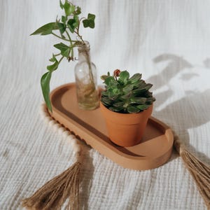 Könnte beinhalten: Ein ovales, terrakottafarbenes Tablett mit gewelltem Rand, verziert mit einer Holzperlengirlande und Jutequasten an beiden Enden. Auf dem Tablett stehen eine kleine, klare Glasflasche mit einem Efeuzweig und ein Terrakottatopf mit einer Sukkulente.