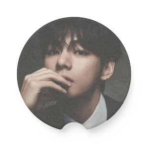 May include: Circular sticker featuring a portrait of a person with dark hair, wearing a dark suit and white shirt. The person is touching their lips with their fingers. The background is a dark gray.