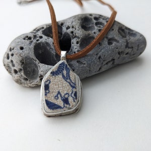 Pode incluir: Colar com pingente artesanal com um fragmento de cerâmica azul e branco em uma moldura prateada, suspenso por um cordão de couro marrom. A cerâmica tem um padrão craquelado e uma ilustração azul.