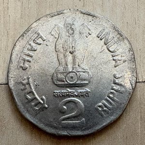 Puede incluir: Una moneda india de plata de 2 rupias. La moneda presenta el emblema del Pilar de Ashoka y el texto "INDIA RUPEE 2". La moneda está ligeramente desgastada, mostrando signos de uso.