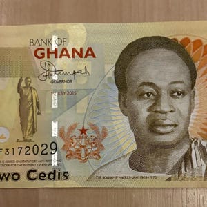 May include: A two-cedi banknote from the Bank of Ghana, featuring a portrait of a man and an illustration of a person. The note has the text "Two Cedis" and the serial number CF3172029. The color scheme is primarily yellow and brown.