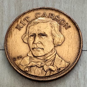 May include: A bronze-colored coin featuring a detailed portrait of Kit Carson. The name "KIT CARSON" is arched across the top. The coin has a textured edge and a slightly worn appearance, suggesting age or use.
