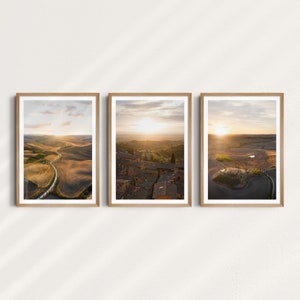 Könnte beinhalten: Set aus drei gerahmten Landschaftsdrucken. Jeder Druck zeigt eine andere Luftaufnahme einer ländlichen Szene bei Sonnenuntergang. Die Szenen umfassen sanfte Hügel, Straßen und Gebäude, die von warmem, goldenem Licht durchflutet sind. Die Rahmen sind aus hellem Holz.