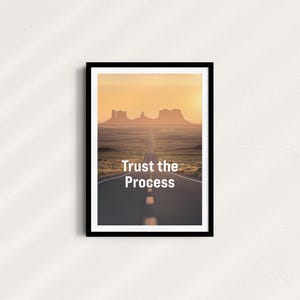 Pode incluir: Uma citação motivacional, "Trust the Process", impressa em um pôster emoldurado em preto com uma foto de uma longa estrada reta que leva a uma cordilheira ao longe. A estrada é ladeada por grama e o céu é de uma cor laranja quente.