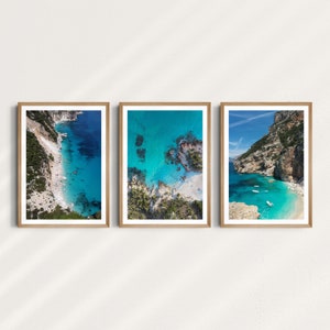 May include: Three framed photographs of aerial views of a turquoise blue ocean with rocky cliffs and beaches. The photos are in wooden frames with white mats.