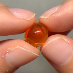 May include: A small, translucent, oval-shaped fire opal gemstone with a vibrant orange hue. The stone exhibits a smooth, polished cabochon surface that catches the light, revealing internal depth and warm, glowing amber tones throughout the gem.