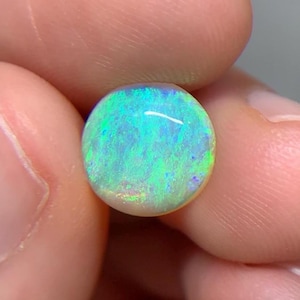 May include: A round, cabochon-cut opal gemstone displaying a vibrant play-of-color with prominent flashes of neon green, electric blue, and subtle hints of yellow across its polished, domed surface, showcasing a high-quality, translucent appearance.