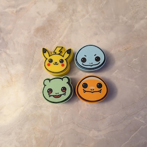 May include: Four colorful character-shaped accessories. The characters include a yellow Pikachu, a blue Squirtle, a green Bulbasaur, and an orange Charmander. Each character has a black outline and a unique facial expression. The accessories are likely for a themed collection.