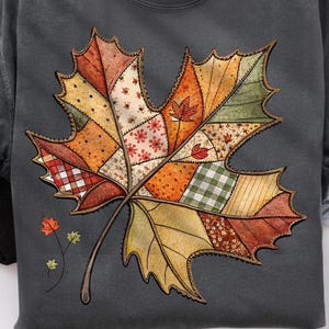 Könnte beinhalten: Dunkelgraues Sweatshirt mit einem großen, farbenfrohen Ahornblatt-Design. Das Blatt besteht aus verschiedenen Patchwork-Mustern in Orange-, Rot-, Grün- und Cremetönen mit einem braunen Stiel. Kleine Blattakzente sind ebenfalls vorhanden.