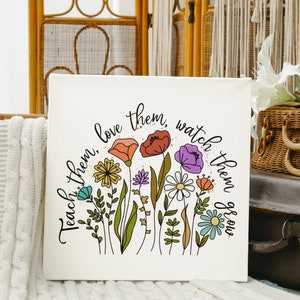 Op de afbeelding: Een witte canvas print met een bloemenontwerp en de tekst "Teach them, love them, watch them grow". De bloemen zijn in tinten roze, paars, geel, oranje en blauw.