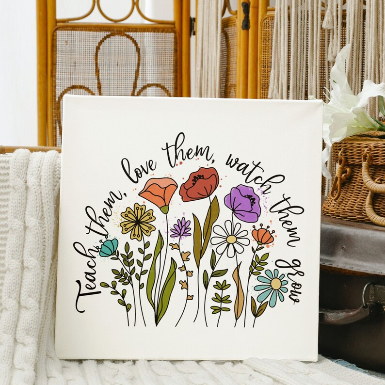 Op de afbeelding: Een witte canvas print met een bloemenontwerp en de tekst "Teach them, love them, watch them grow". De bloemen zijn in tinten roze, paars, geel, oranje en blauw.