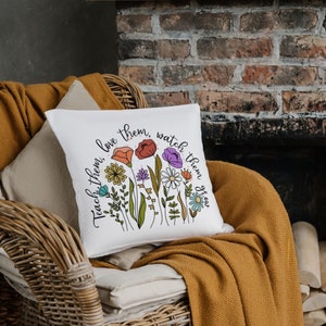 Op de afbeelding: Een wit kussen met een bloemenpatroon en de tekst "Teach them, love them, watch them grow".