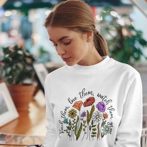 Op de afbeelding: Witte trui met een bloemenontwerp en de tekst "Teach them, love them, watch them grow."