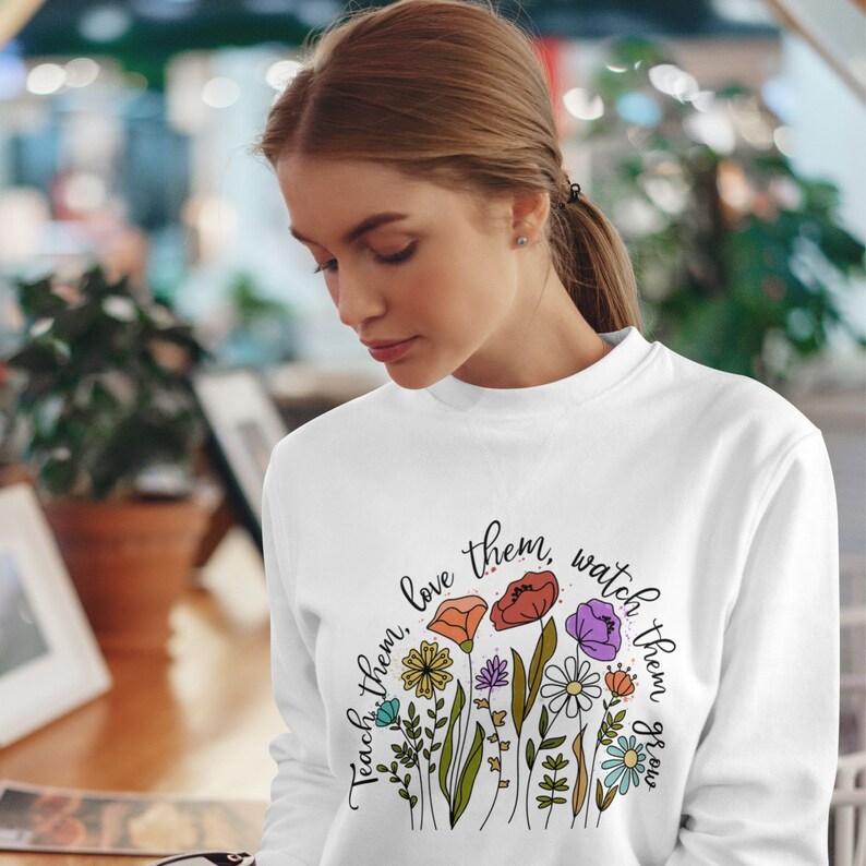 Op de afbeelding: Witte trui met een bloemenontwerp en de tekst "Teach them, love them, watch them grow."