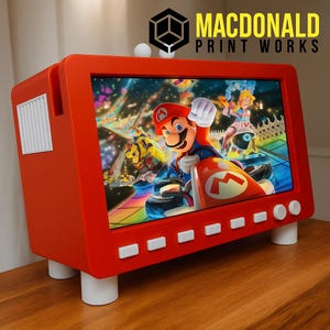 Puede incluir: Un televisor rojo de estilo retro con una pantalla que muestra una escena de carreras de Mario Kart. El televisor tiene botones y patas blancas, y el texto "MACDONALD PRINT WORKS" es visible en la parte superior derecha.