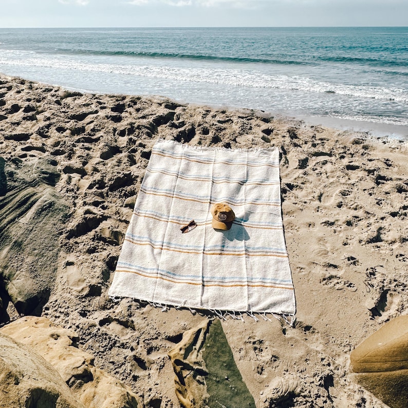 Beach blanket made from upcycled cotton, perfect for beach days, picnics, or yoga. This colorful Mexican blanket is a versatile outdoor blanket, ideal for summer adventures, camping, or as a cozy home decor throw. Soft, lightweight, and eco-friendly, it doubles as a boho throw blanket, blanket gift, or decorative layer for any coastal space. Handwoven by artisans, this sustainable, upcycled cotton blanket adds comfort and style wherever it goes.