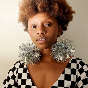 May include: A person with brown curly hair wearing a black and white checked shirt with large silver sparkly earrings.