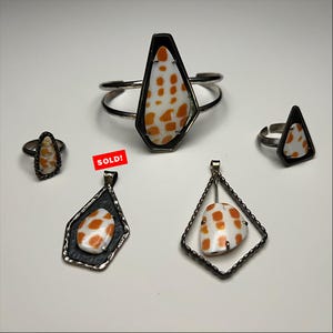 May include: A collection of jewelry featuring white and orange spotted shell inlays set in dark metal frames. The set includes a cuff bracelet, two rings, and two pendants, one of which has a red rectangular tag with the text SOLD! printed on it.