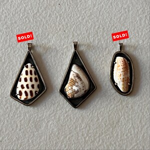 May include: Three silver-toned pendants with natural seashells. The pendants have a geometric design, with the shells encased in a dark metal frame. One pendant has a shell with brown spots, another with tan stripes, and the third with brown spots. The word "SOLD!" is on two of the pendants.