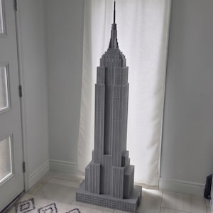 May include: A large, grey, three-dimensional model of the Empire State Building featuring a stepped architectural design, a central tower, and a pointed spire at the top. The structure displays a grid-like pattern representing windows and floors.