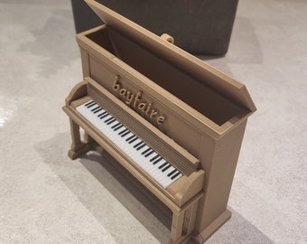 Peut inclure: Un petit piano en bois brun avec un clavier blanc et le mot "bayfaire" inscrit sur le dessus. Le piano est ouvert et le couvercle est relevé.